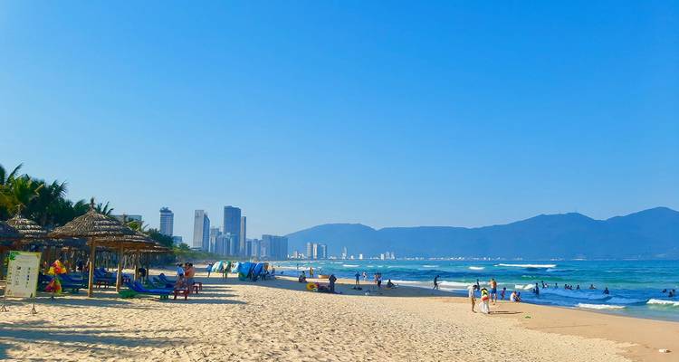 Playa soleada en la ciudad de Da Nang con sombrillas, bañistas y un horizonte de rascacielos a lo lejos respaldado por montañas.
