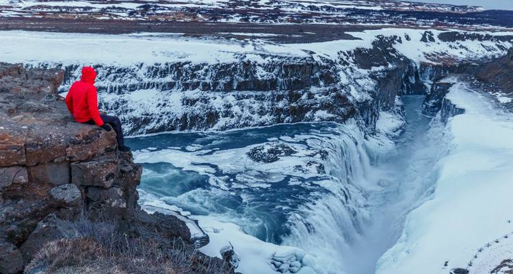 Person sitting on cliff edge overlooking icy waterfall.