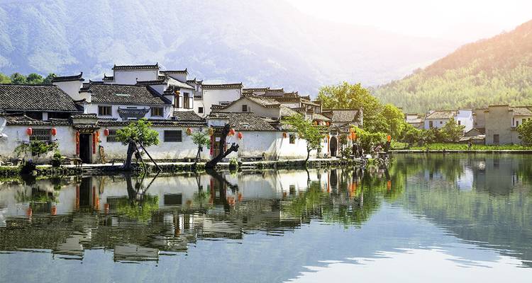 Village chinois traditionnel au bord d'un étang avec des montagnes en arrière-plan.