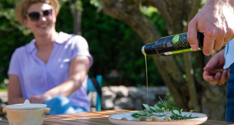 Gros plan d'huile d'olive versée sur une assiette d'olives et d'herbes lors d'une dégustation en plein air, avec un invité souriant flou en arrière-plan.