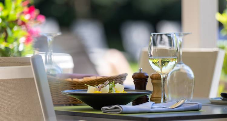 Table de restaurant élégante en plein air dressée avec du vin blanc, une mise en bouche gastronomique, une corbeille à pain et des fleurs colorées en arrière-plan légèrement flou.