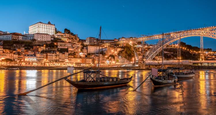 A scenic night view of Porto, Portugal, featuring boats on the river and the illuminated Dom Luís I Bridge.
