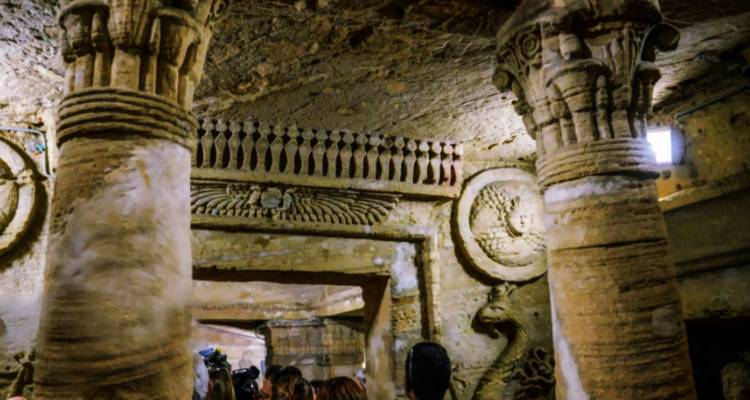 Intérieur d'une tombe égyptienne antique avec des colonnes sculptées et des personnes qui explorent.