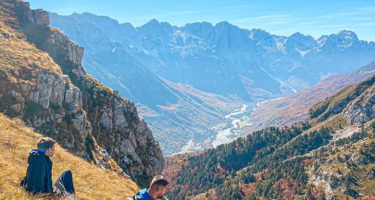 Deux personnes assises sur une colline herbeuse surplombant une vallée avec des montagnes au loin.