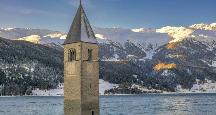 Gezonken kerktoren in een meer met besneeuwde bergen op de achtergrond.