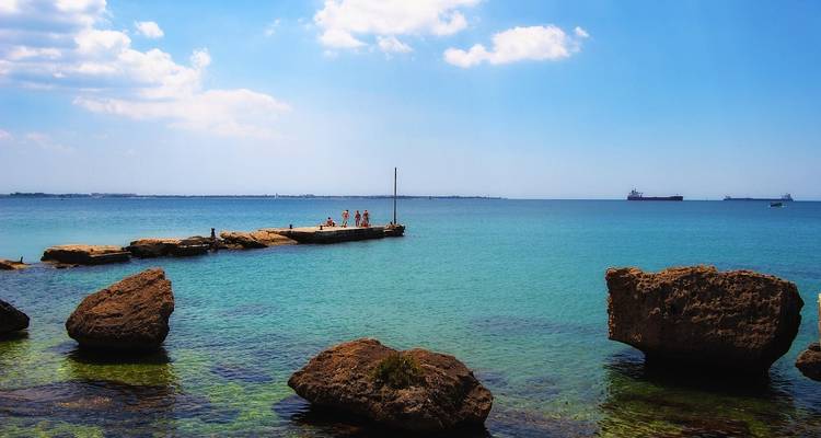 Mer bleue limpide avec une jetée en pierre et des navires au loin.