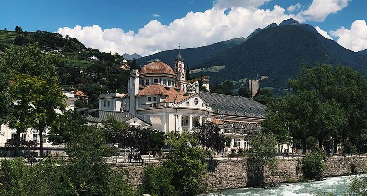 Historic building by a river with mountains in the background.