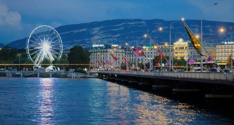 Scène en soirée au bord de l'eau avec une grande roue illuminée, les lumières du pont se reflétant sur le lac avec les montagnes en arrière-plan.