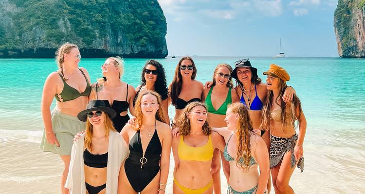 Group of women in swimsuits posing on a beach with clear water and cliffs.