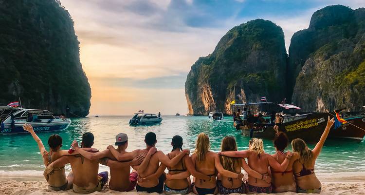 A group of people sitting on the beach with arms around each other, overlooking the sea and boats.
