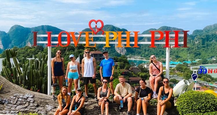 People posing in front of a sign that reads 'I Love Phi Phi' with scenic hills and water in the background.