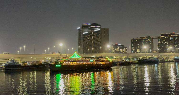 A dhow boat decorated with green lights, near a city skyline at night.