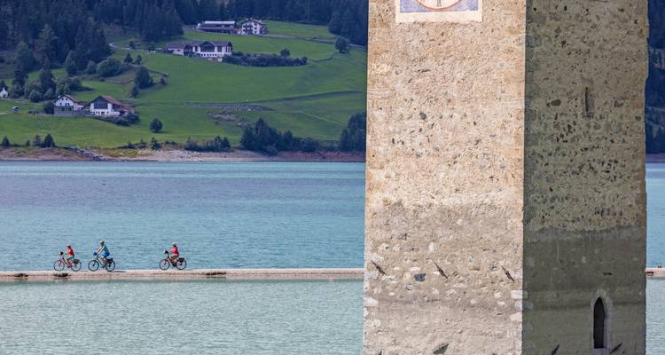 Radfahrer fahren entlang des Damms des Resia-Sees mit dem berühmten versunkenen Kirchturm im Vordergrund