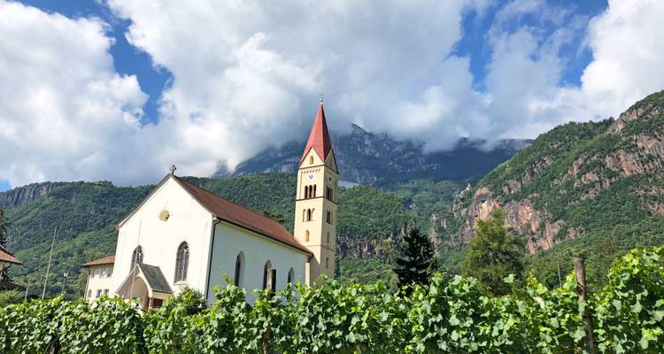 A charming white church with a red steeple stands among green vineyards, backed by forested mountains partly shrouded in clouds under a bright sky.