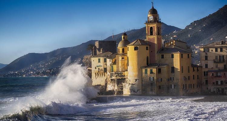 Waves crashing against a coastal church with rugged mountains behind.