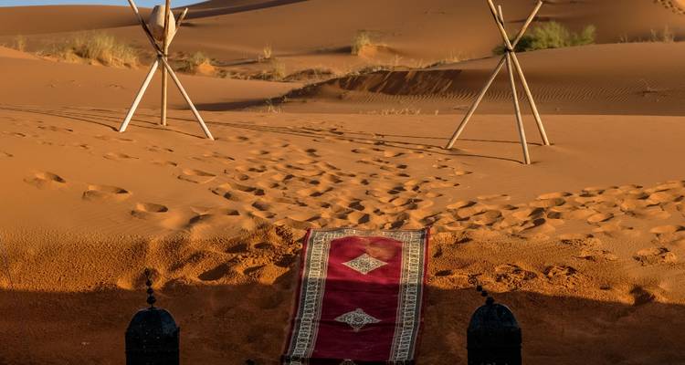A red carpet leads into golden desert dunes beside simple wooden tripod poles at a camp.