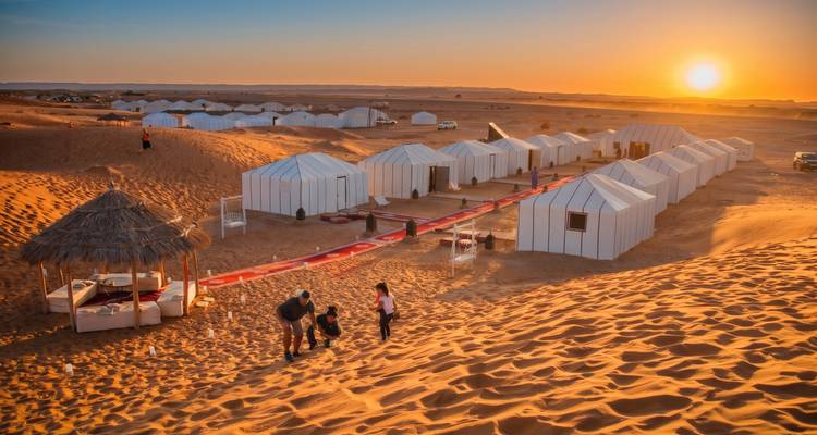Luxury white desert camp tents line a red carpet on golden dunes at sunset in the Sahara.