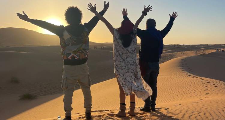 Trois personnes levant les bras en se tenant debout sur des dunes de sable au coucher du soleil.