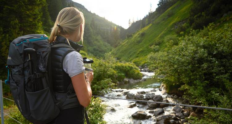Frau mit einem Rucksack, die einen Gebirgsbach in einem grünen Tal betrachtet.