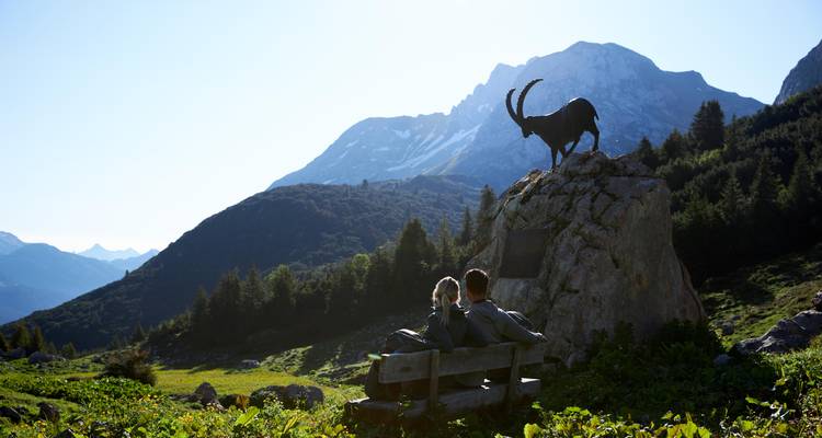 Paar sitzt auf einer Bank mit einer Bergziegen-Skulptur in einer Berglandschaft.