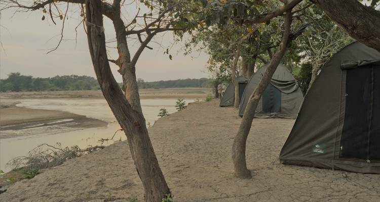 Campamento sencillo a orillas del río con carpas de lona oscura instaladas en tierra agrietada junto a un río sinuoso.
