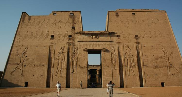 Temple ancien avec des murs sculptés sous un ciel dégagé, avec des touristes marchant devant.