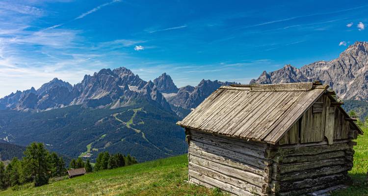 Cabane en bois sur une colline herbeuse avec une chaîne de montagnes en arrière-plan.
