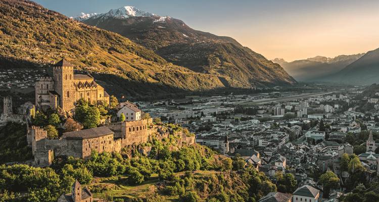 Vue aérienne d'une ville avec un château et un décor de montagnes au coucher du soleil.