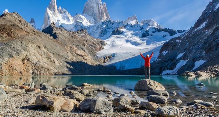 Person raising arms standing on a rock in front of snowy mountains.