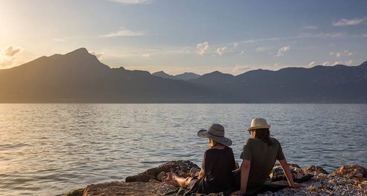 Deux personnes assises sur des rochers au bord du lac de Garde, profitant de la vue du coucher de soleil sur les montagnes.