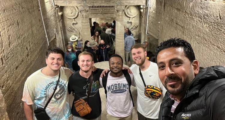 Grupo de turistas posando para una selfie dentro de antiguas catacumbas de piedra con tallados ornamentados.