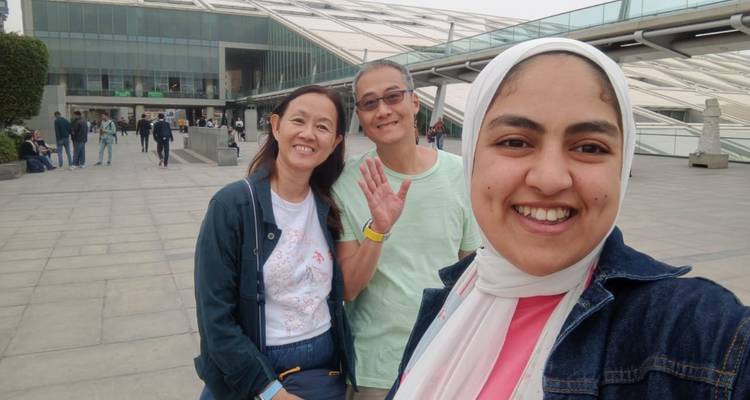Turistas sonriendo fuera de la plaza de la Bibliotheca Alexandrina en un día nublado.