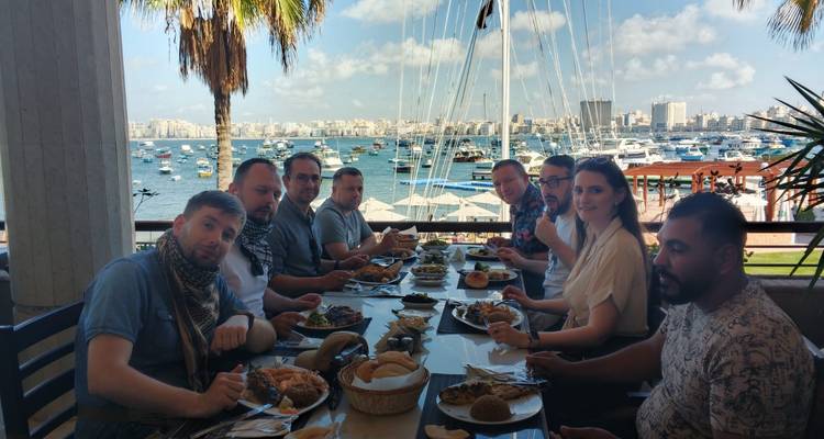 Gran mesa de viajeros cenando en una terraza con vistas al puerto de Alejandría lleno de botes.