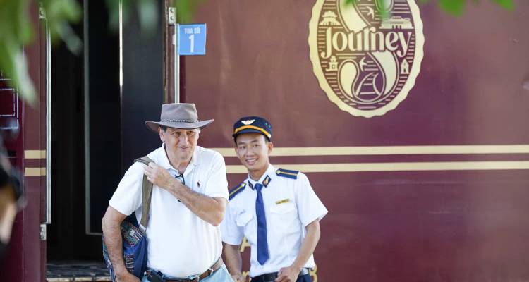 Un homme avec un chapeau et un contrôleur de train souriant à l'extérieur d'un wagon de train avec le logo 'The Journey'.