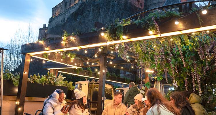 Grupo de amigos cenando bajo luces centelleantes con imponente castillo de piedra alzándose sobre ellos