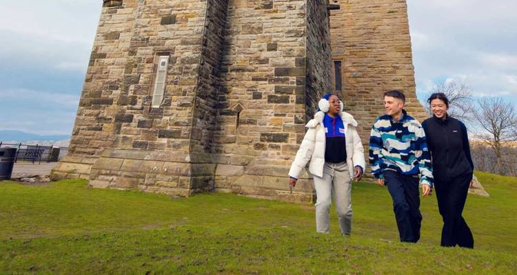 Three friends stroll past an old stone tower set on a grassy hill with winter light.