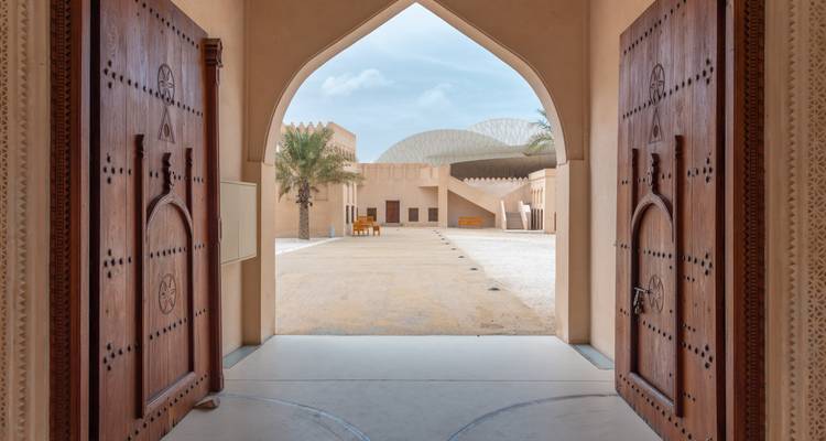 Vue à travers des portes traditionnelles en bois sur une cour avec des palmiers et une architecture moderne.