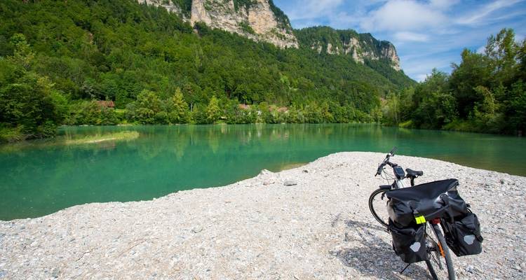 Bicicleta en la orilla de un lago con montañas y árboles al fondo.