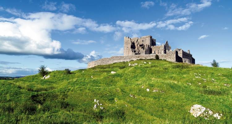 Eine einsame mittelalterliche Steinburg steht auf einem leuchtend grünen Hügel unter einem strahlend blauen Himmel mit vereinzelten Wolken.