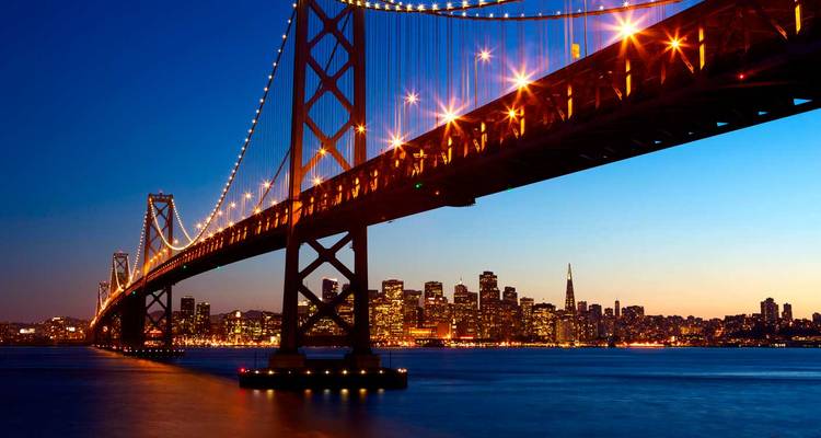 San Francisco–Oakland Bay Bridge lit at twilight with city skyline glowing across calm bay waters