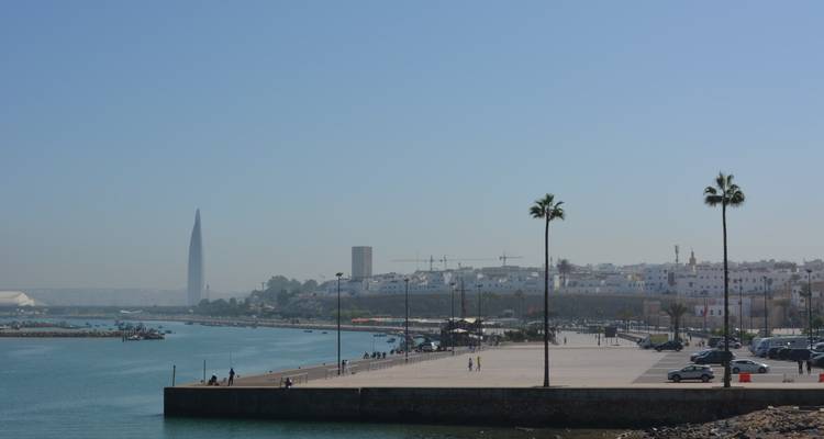 Breite Pier, die sich in ruhige Gewässer erstreckt mit vereinzelten Besuchern, zwei Palmen und Rabats moderner Skyline in diesigem Licht.