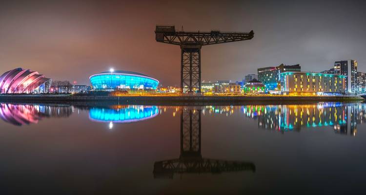 Vue panoramique d'un paysage urbain la nuit avec des bâtiments illuminés et une rivière reflétant les lumières.