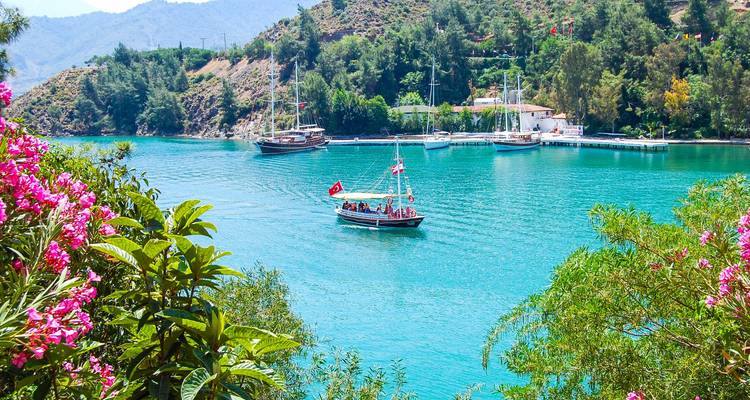 A small boat on a turquoise river bordered by lush hills and blooming flowers.