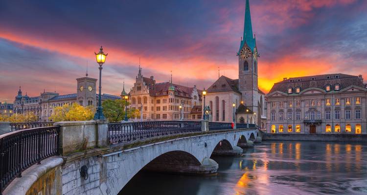 Puente de piedra sobre un río reflectante que lleva a una torre de iglesia histórica bajo un cielo dramático de atardecer en una ciudad europea.
