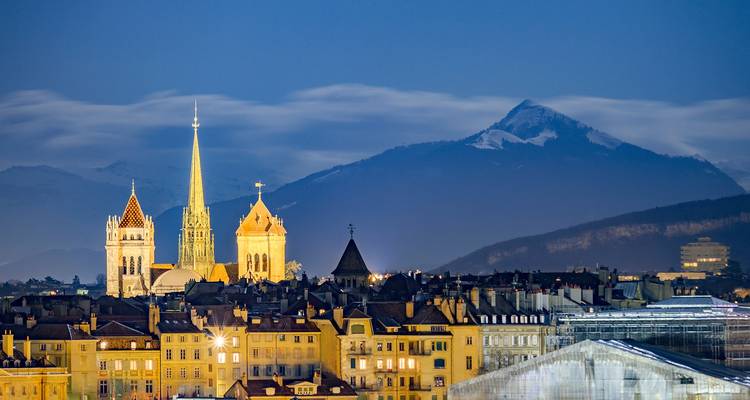 Horizonte vespertino con agujas de catedral iluminadas contra montañas nevadas más allá de una ciudad europea histórica.