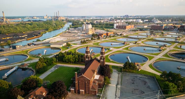 Vue aérienne d'un front de rivière industriel avec des bassins de décantation circulaires et une église en brique au premier plan, paysage urbain étendu au-delà.