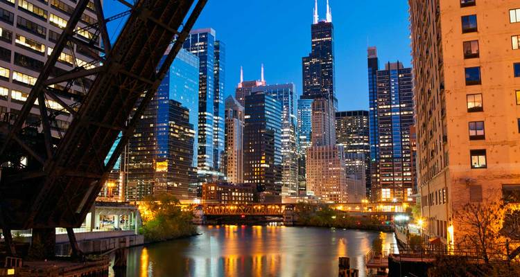 Evening view of Chicago’s illuminated skyline reflecting on the river with an open steel bridge in the foreground.