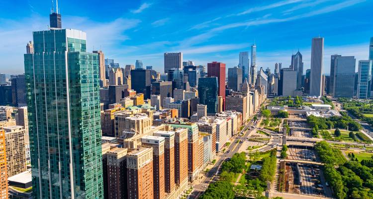 Bright daytime aerial of Chicago’s downtown towers, parkland, and Lake Michigan shoreline under blue skies.