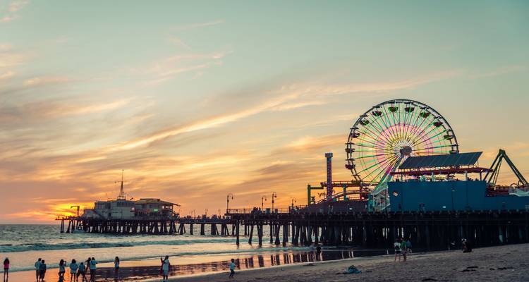 Golden sunset at Santa Monica Pier with illuminated Ferris wheel, beachgoers and gentle surf.