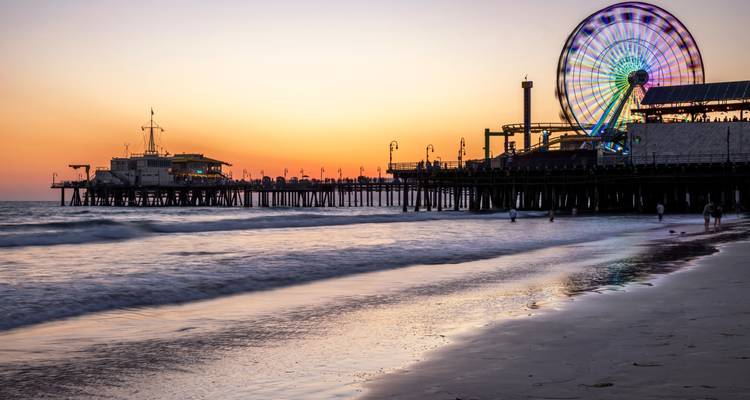 After-sunset glow over Santa Monica Pier with neon Ferris wheel and gentle surf lapping the beach.
